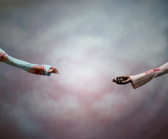 Two arms reach toward each other with open hands against a backdrop of diffused pink, white, and grey clouds, creating an ethereal scene.