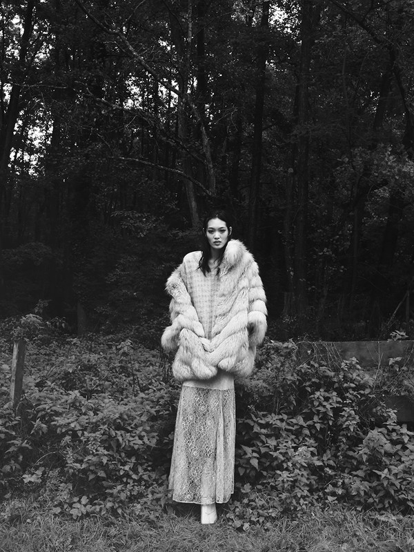 A long shot of a woman in a forest wearing a fur coat and a long skirt, posing for a fashion editorial with a slightly serious expression.