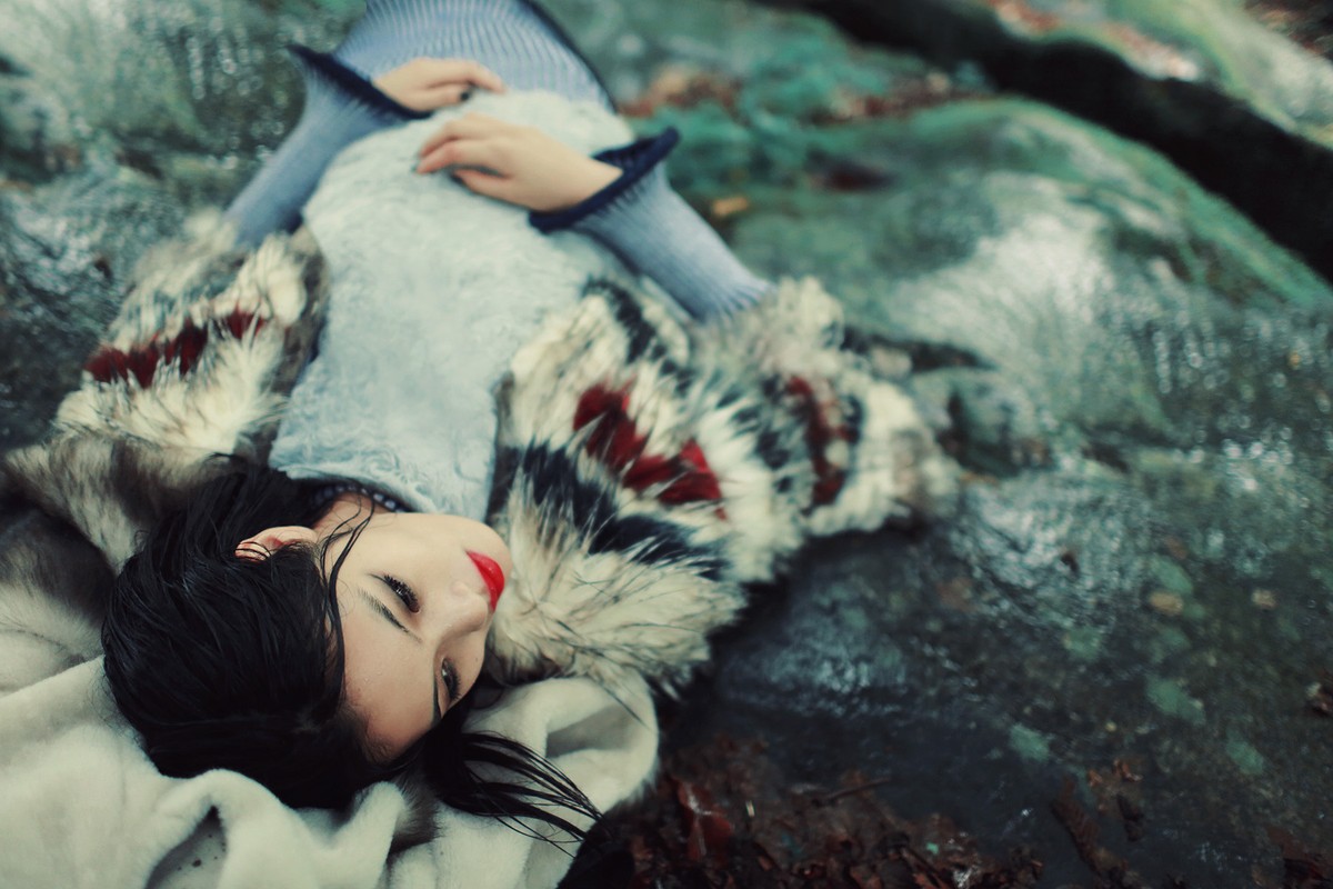 Woman wearing patterned faux fur coat lying on damp mossy rocks near shallow stream water.