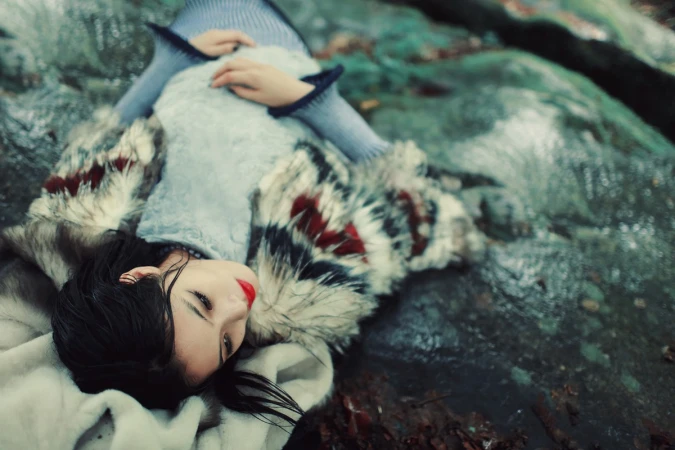 Woman wearing patterned faux fur coat lying on damp mossy rocks near shallow stream water.