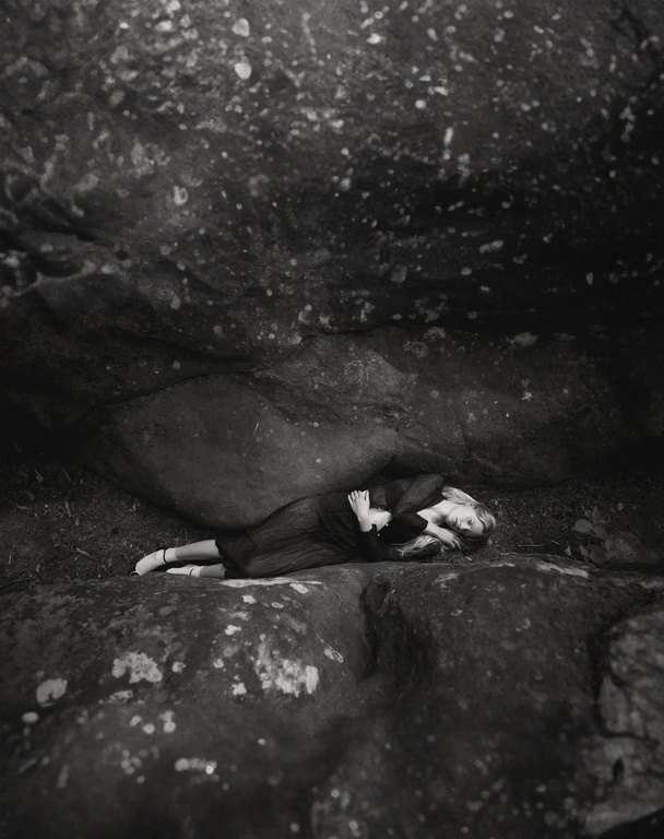 Monochrome portrait of a woman lying down on a rock outdoors, with a rock overhang above, creating a dramatic and intimate scene.