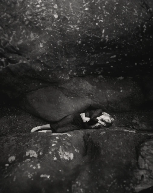 Monochrome portrait of a woman lying down on a rock outdoors, with a rock overhang above, creating a dramatic and intimate scene.