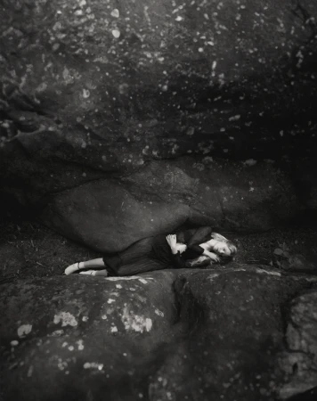 Monochrome portrait of a woman lying down on a rock outdoors, with a rock overhang above, creating a dramatic and intimate scene.