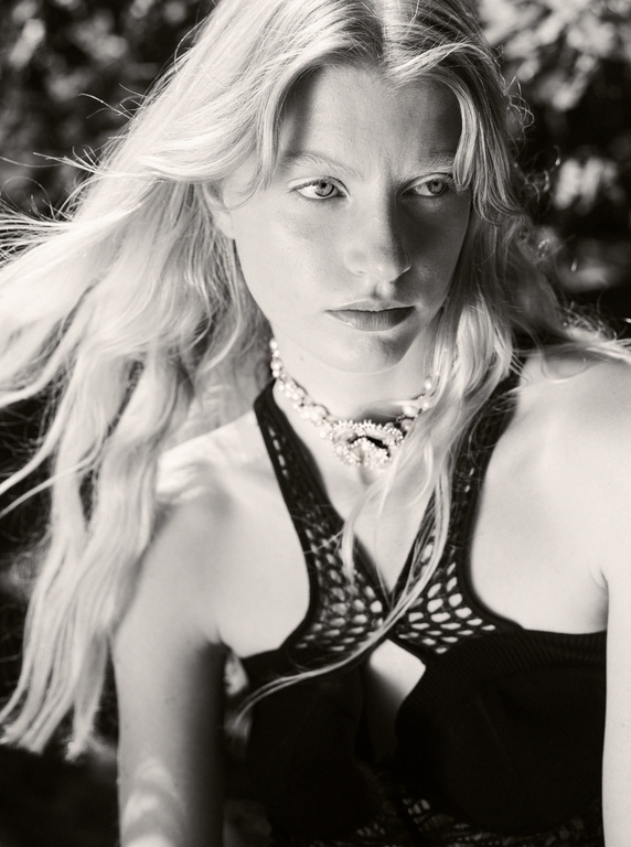 Monochromatic portrait of an adult woman with long blonde hair wearing a dark, open-weave top and an ornate pearl choker. Outdoor setting.