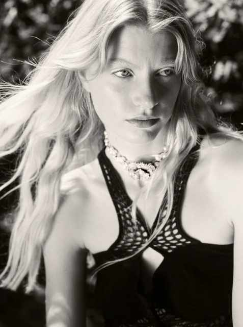 Monochromatic portrait of an adult woman with long blonde hair wearing a dark, open-weave top and an ornate pearl choker. Outdoor setting.