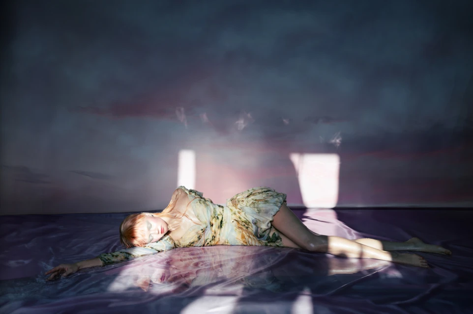 Woman in a floral dress lies on a reflective surface in front of a wall with light flares, creating an ethereal, dreamlike fashion scene.