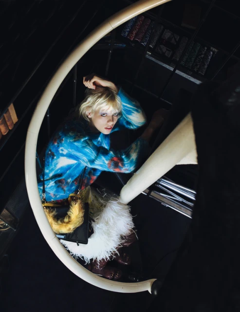 Woman in tie-dye top poses on spiral staircase. The indoor shot includes a small handbag, white furry trim, and dark contrasting background.