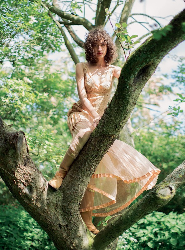 Woman with curly brown hair in a peach dress perched among tree branches in a forest by photographer Alexandra Sophie