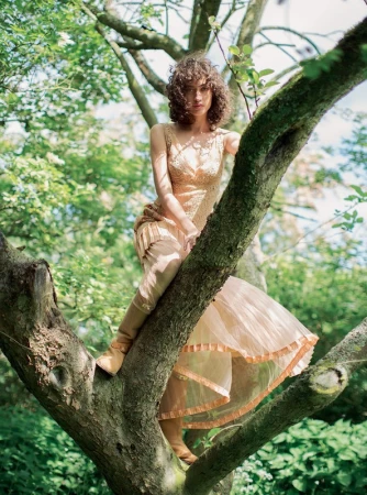 Woman with curly brown hair in a peach dress perched among tree branches in a forest by photographer Alexandra Sophie