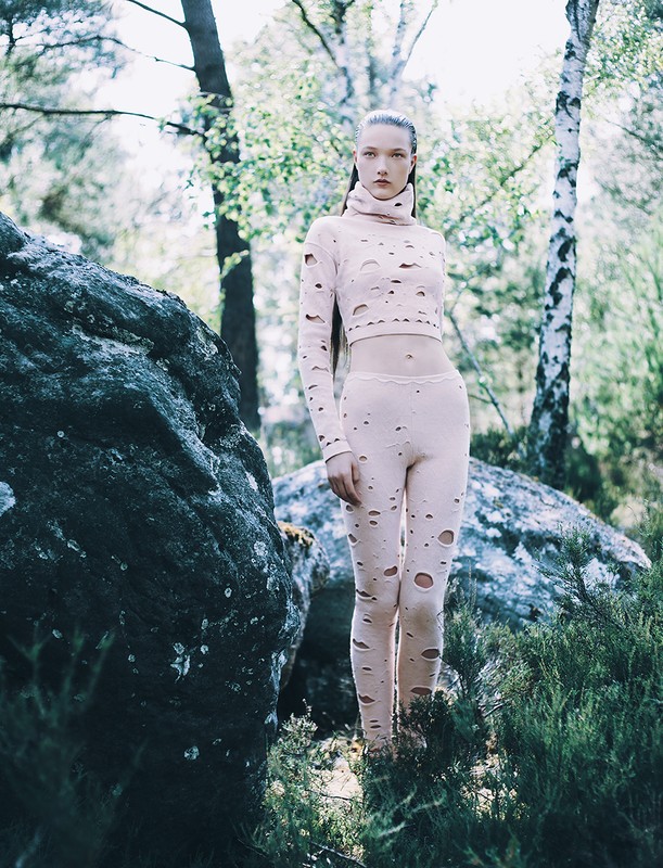 A slender woman in a light pink, distressed long-sleeved crop top and leggings stands in a forest with rocks and scattered sunlight.
