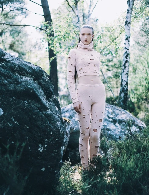 A slender woman in a light pink, distressed long-sleeved crop top and leggings stands in a forest with rocks and scattered sunlight.