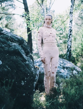 A slender woman in a light pink, distressed long-sleeved crop top and leggings stands in a forest with rocks and scattered sunlight.
