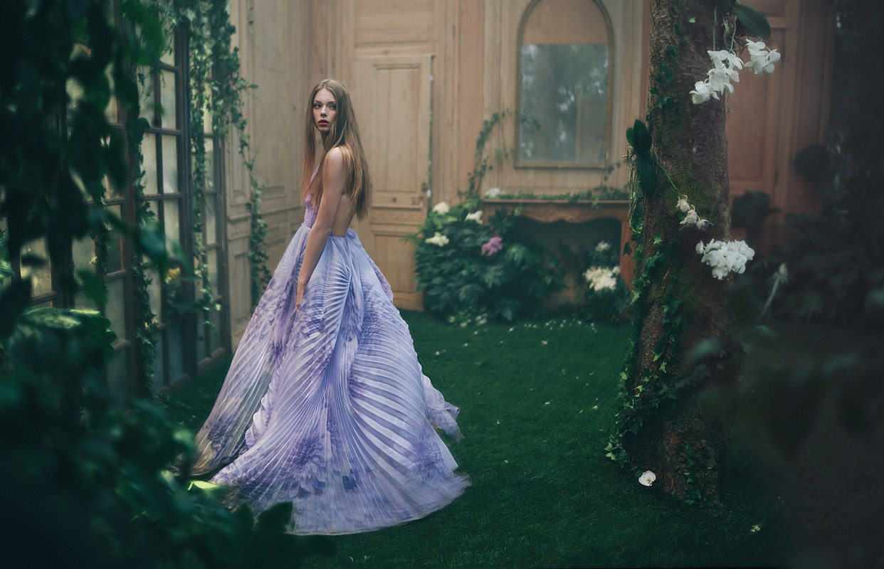 Woman in flowing lavender gown stands in a garden-like room with a tree covered in white flowers. Interior fashion shoot.