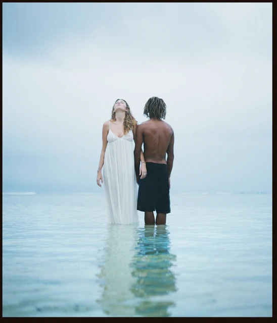 A couple stands waist-deep in clear ocean water. The woman wears a white dress and looks upward; the man has dreadlocks and wears black swim trunks.