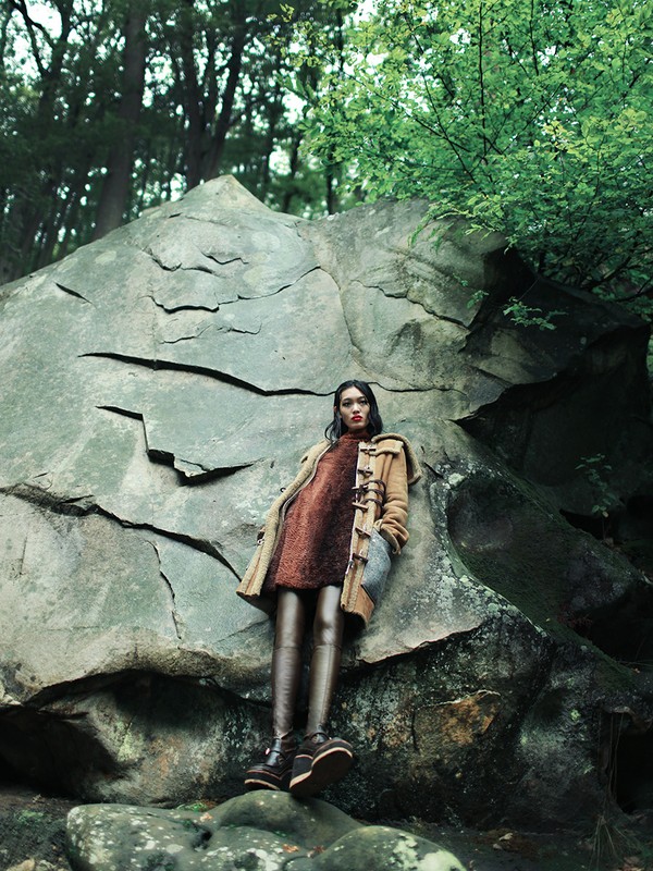 A woman in a coat poses next to a large boulder in a forest. She is wearing a dress, tights, and boots in a fashion shot.