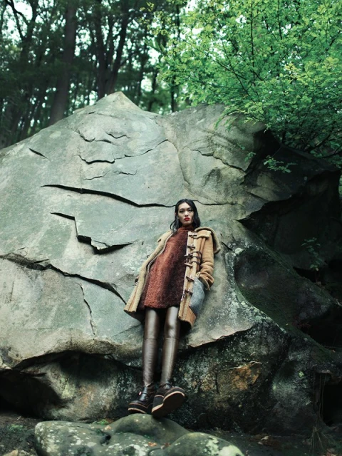 A woman in a coat poses next to a large boulder in a forest. She is wearing a dress, tights, and boots in a fashion shot.