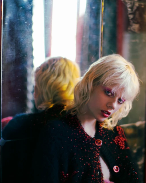 Blonde woman wearing a black and red beaded jacket poses in front of a mirror, reflecting her head and shoulders in a blurred image.