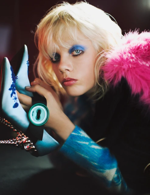 Fashion shot of a woman with blonde hair and blue eye shadow holding a pair of light blue heels with flower details and a pink boa.