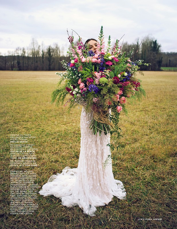 Woman in lace dress holding a large floral bouquet in a field, showcasing fashion and floral design against a natural outdoor backdrop.
