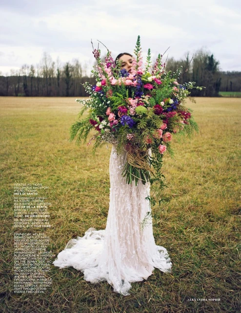 Woman in lace dress holding a large floral bouquet in a field, showcasing fashion and floral design against a natural outdoor backdrop.