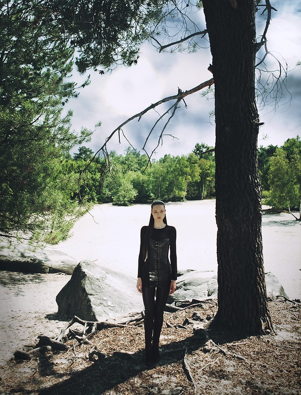 Adult female fashion model in a black outfit stands near a tall tree within a forest environment, bathed in natural daylight.