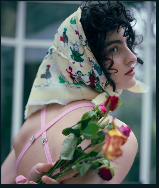 Woman with curly hair poses wearing printed headscarf, pink bra straps, holding roses in front of a greenhouse window. Close-up portrait.