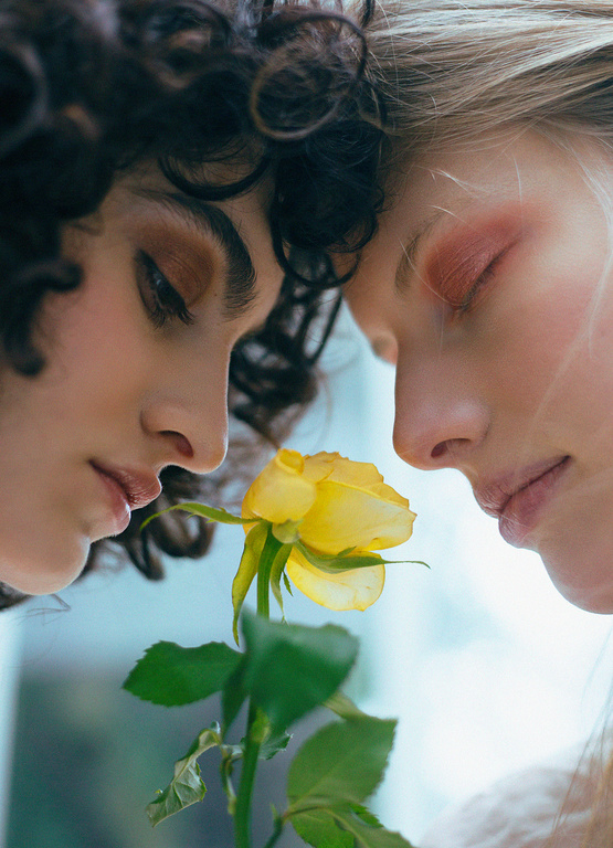 Close-up of two women with eyes closed, heads together, near a yellow rose. Beauty shot with soft lighting and focus on skin and flower.