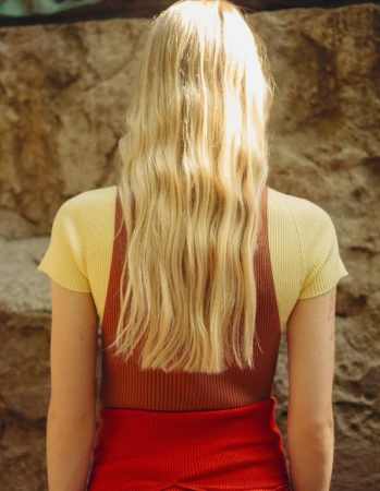 Rear view of a blonde woman wearing a yellow top and brown knit vest against a stone wall.