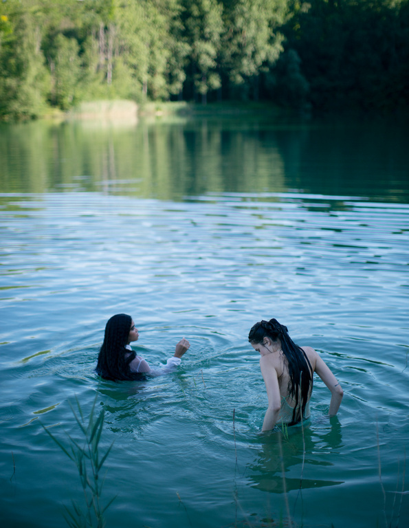 Two women swimming in a calm forest lake, captured from behind in soft natural light by photographer Alexandra Sophie.
