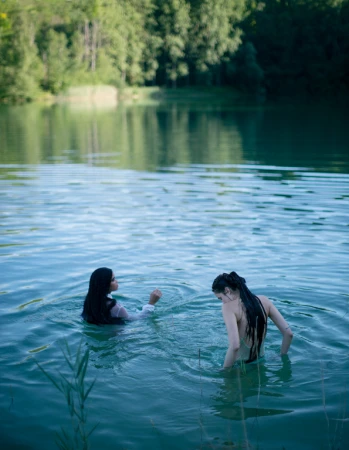 Two women swimming in a calm forest lake, captured from behind in soft natural light by photographer Alexandra Sophie.