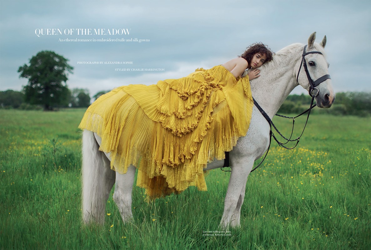 A woman in a yellow gown rests on a white horse in a meadow setting, evoking a serene and fashion-forward outdoor scene.