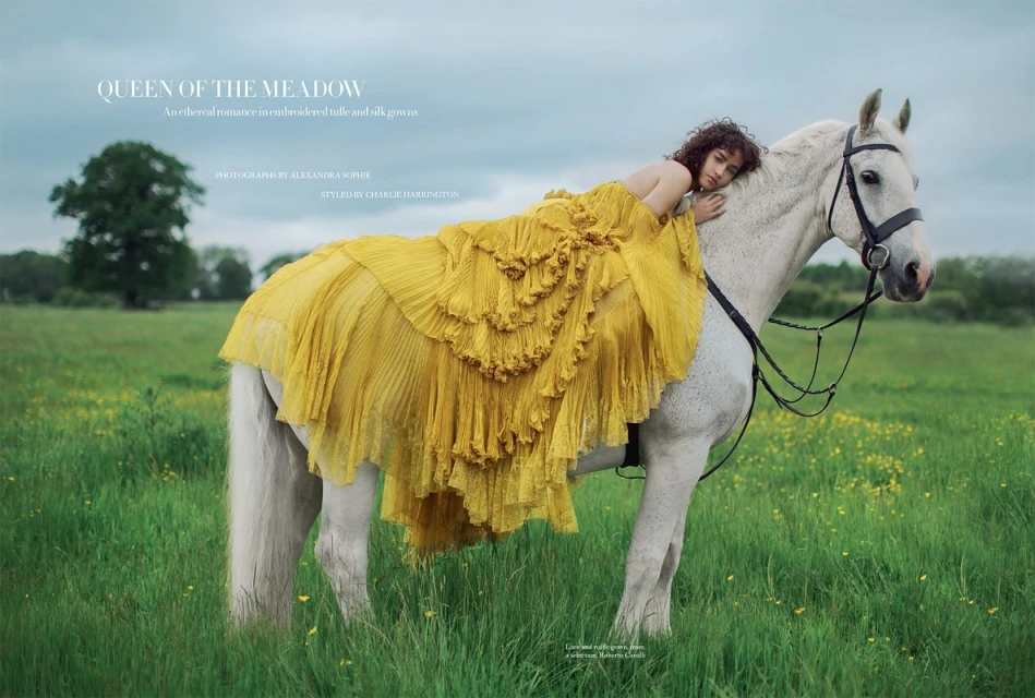 A woman in a yellow gown rests on a white horse in a meadow setting, evoking a serene and fashion-forward outdoor scene.
