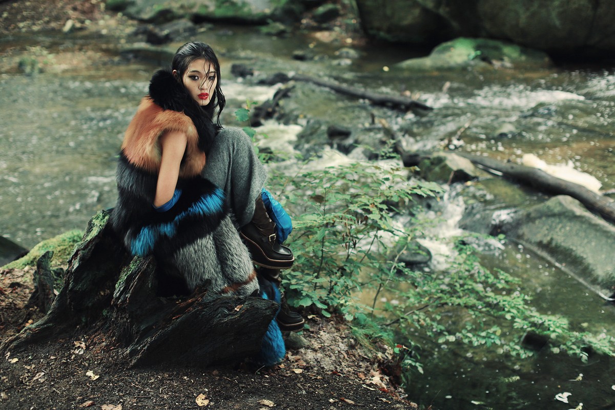Woman in fur-trimmed coat and boots poses in the woods next to a flowing stream, creating a nature-inspired fashion editorial shot.