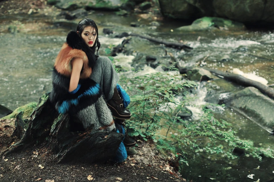 Woman in fur-trimmed coat and boots poses in the woods next to a flowing stream, creating a nature-inspired fashion editorial shot.