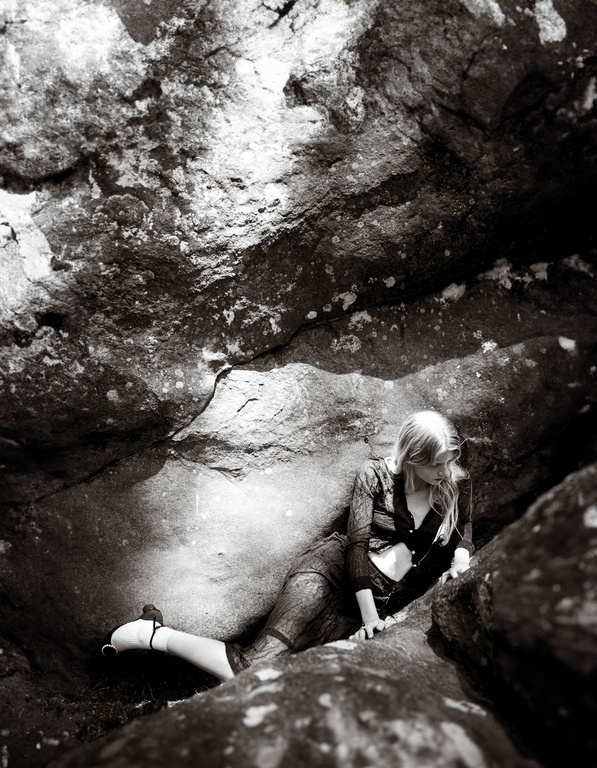 A monochrome image shows a woman with blonde hair lying on rocks, with large rock formations surrounding her in an outdoor environment.