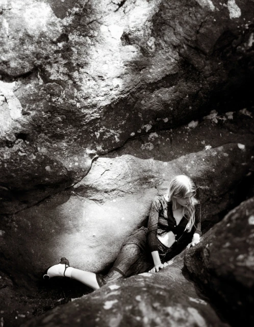 A monochrome image shows a woman with blonde hair lying on rocks, with large rock formations surrounding her in an outdoor environment.