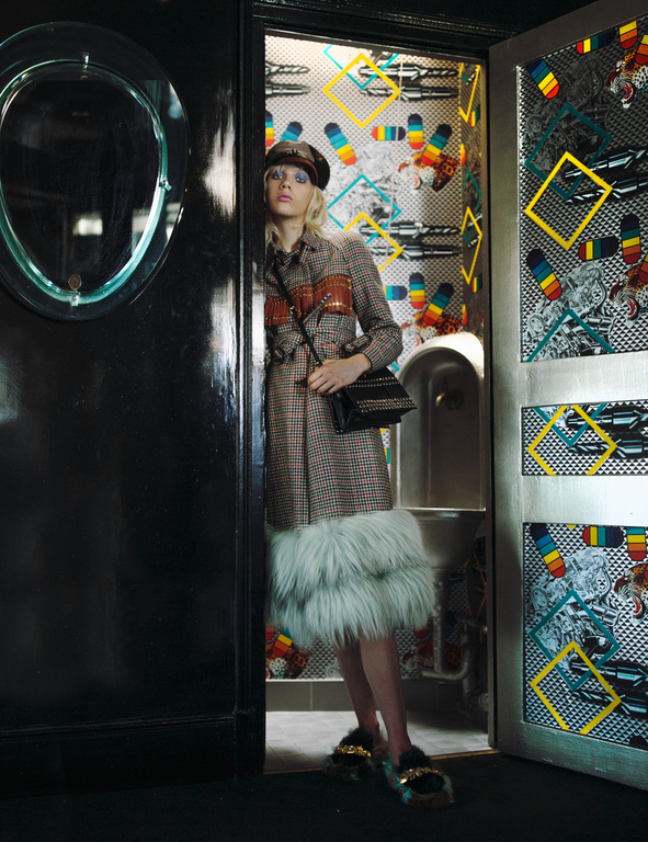 A stylish woman with blond hair and a beret poses in a doorway wearing a patterned coat and fuzzy shoes in an indoor setting.