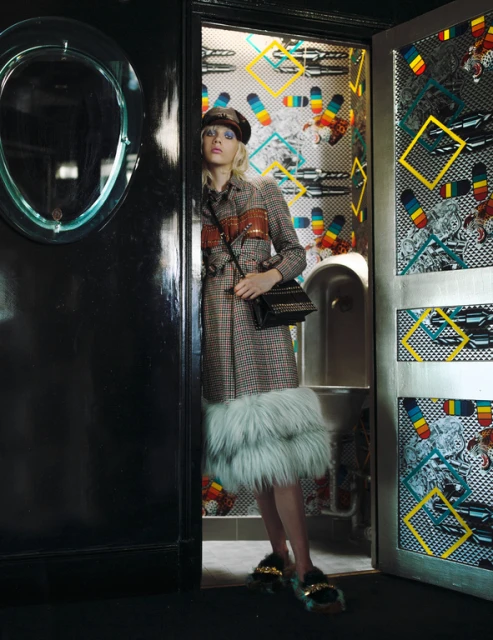 A stylish woman with blond hair and a beret poses in a doorway wearing a patterned coat and fuzzy shoes in an indoor setting.