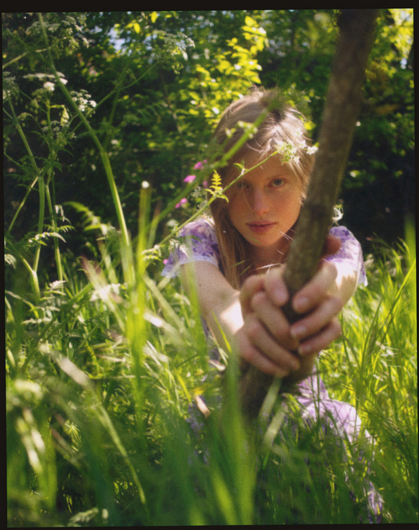 Young woman holding a vertical tree branch amidst tall green grass and wildflowers in a natural outdoor setting