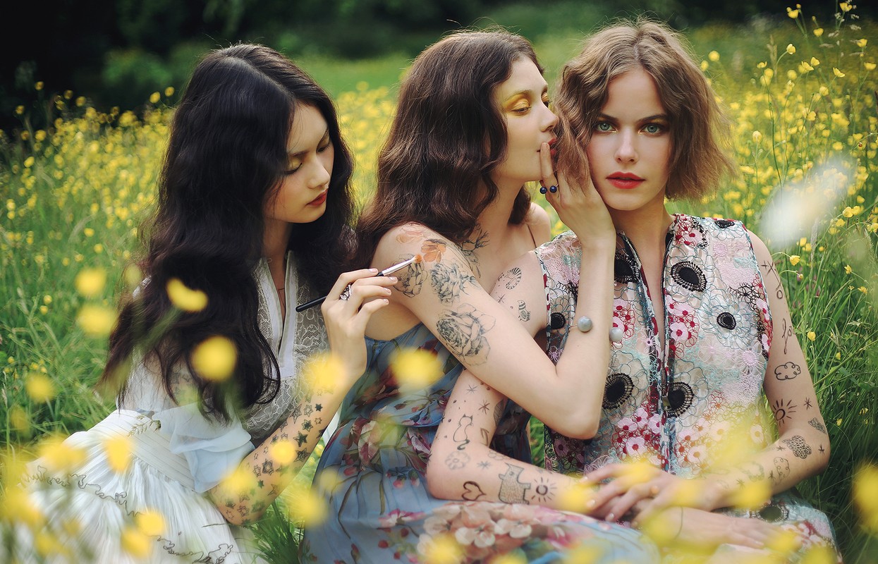 Three women pose in a sunlit flower field, with tattoos, floral dresses, and makeup, evoking a vintage, ethereal aesthetic.