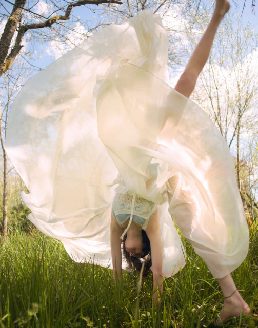 Young woman in a light dress balances in a handstand in a grassy field, trees and sky visible in background during daytime.