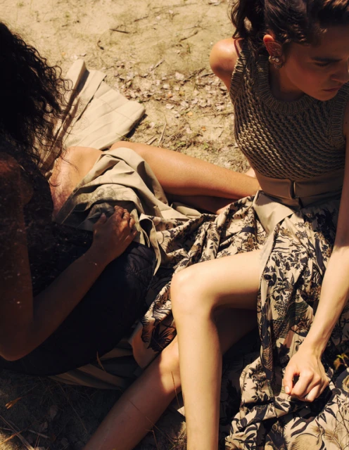 Two women pose in a sunlit outdoor setting, one wearing a tan knit top and floral skirt, the other in dark top and patterned skirt.