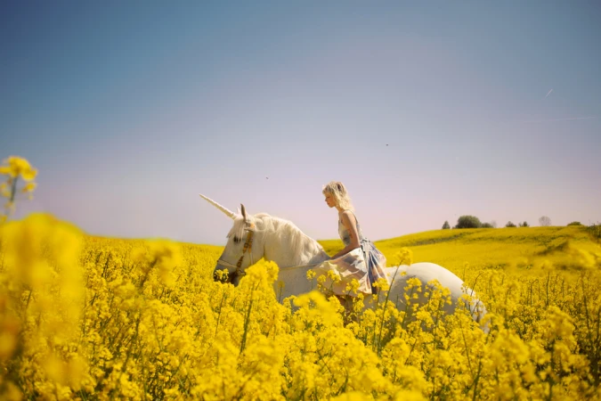 Woman in flowing dress rides white horse with unicorn horn through field of yellow rapeseed flowers under a clear blue sky.