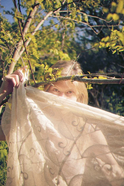 Woman in nature partially concealed by patterned fabric, framed by tree branches. Sunlight filters through the green foliage.