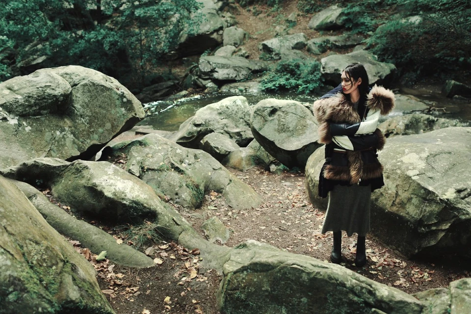 Woman in a fur-trimmed jacket standing among moss-covered rocks near a stream in a wooded area. Fashion editorial with natural environment.