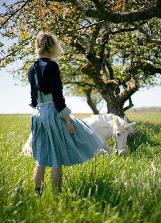 Woman in blue skirt stands in a grassy field by a white horse and a flowering tree, bathed in soft daylight.