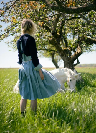 Woman in blue skirt stands in a grassy field by a white horse and a flowering tree, bathed in soft daylight.