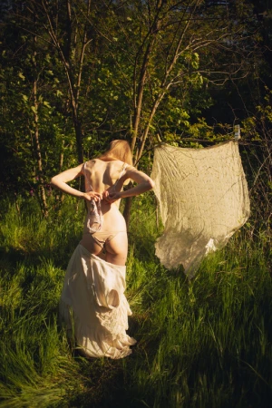 Blonde woman seen from behind adjusting her underwear while standing in a lush green forest clearing during sunset.