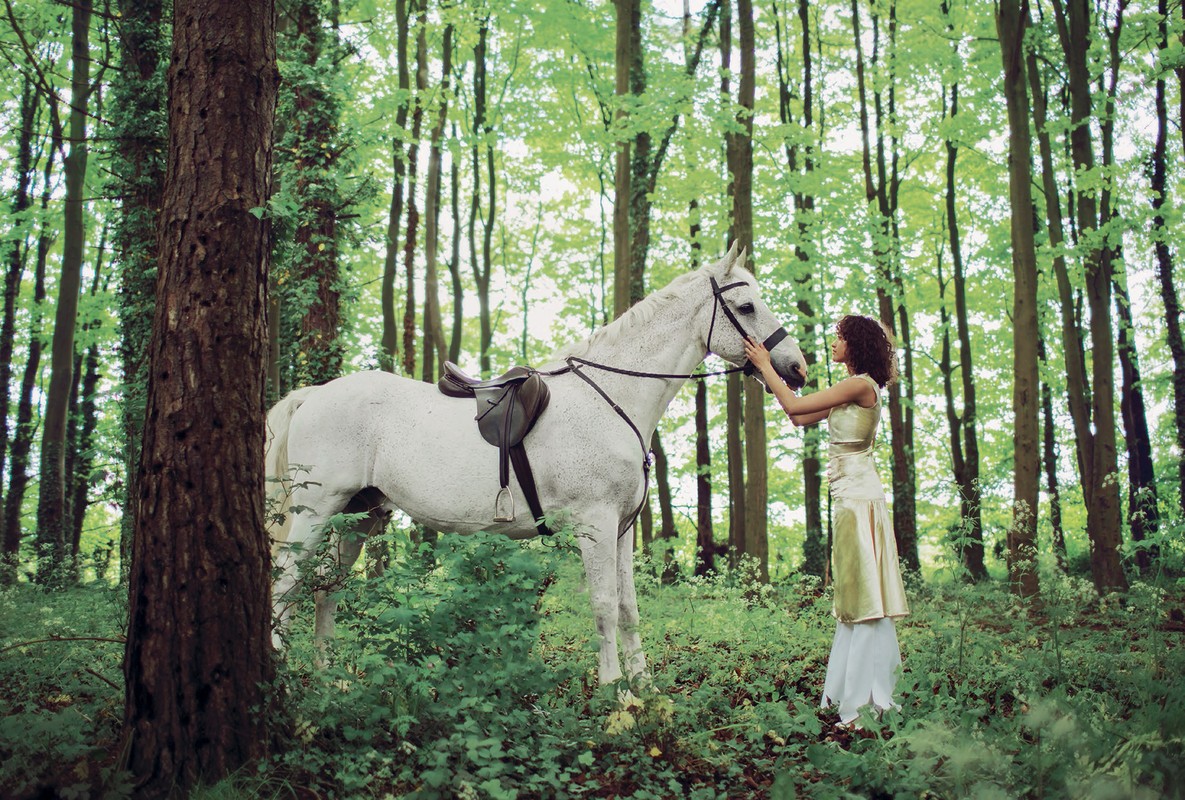 A woman in a gold dress stands in a lush forest beside a saddled white horse, gently touching its face.