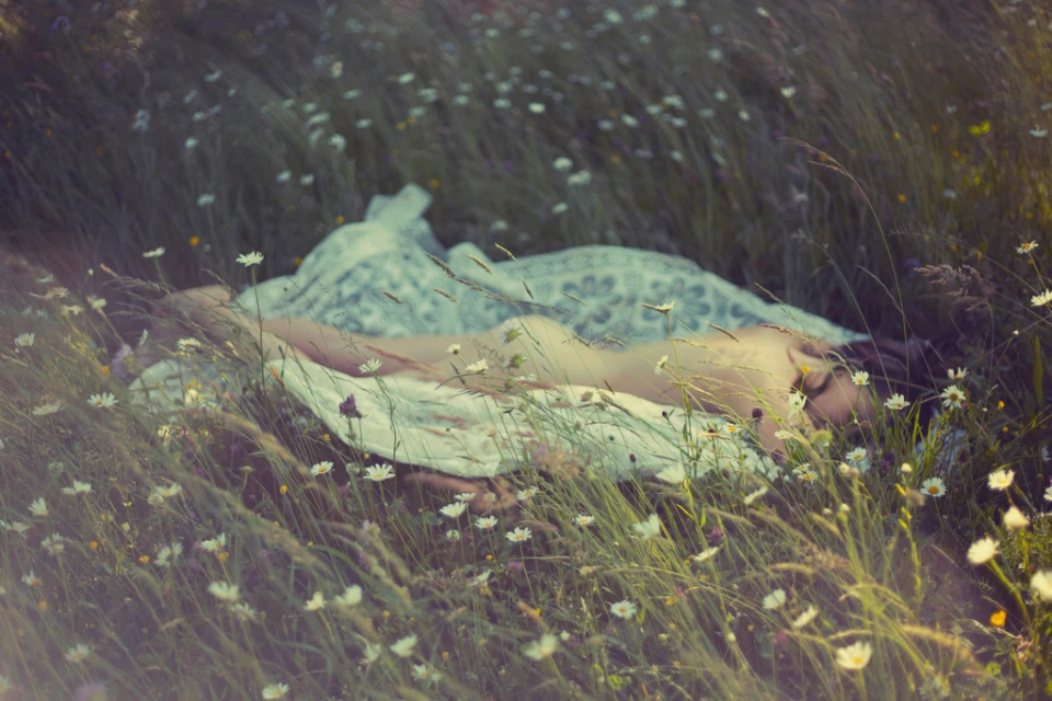 Young woman lies in a meadow of daisies in soft light, wearing a white dress and blanket, with an eye-level, outdoor view.